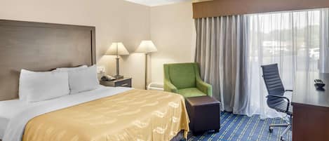 Una habitación de hotel con una cama grande, dos lámparas de noche, un sillón verde, una silla de escritorio negra y un ventanal con cortinas.