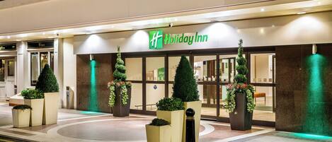 A Holiday Inn hotel entrance with a modern design, featuring large planters with greenery and a welcoming sign.