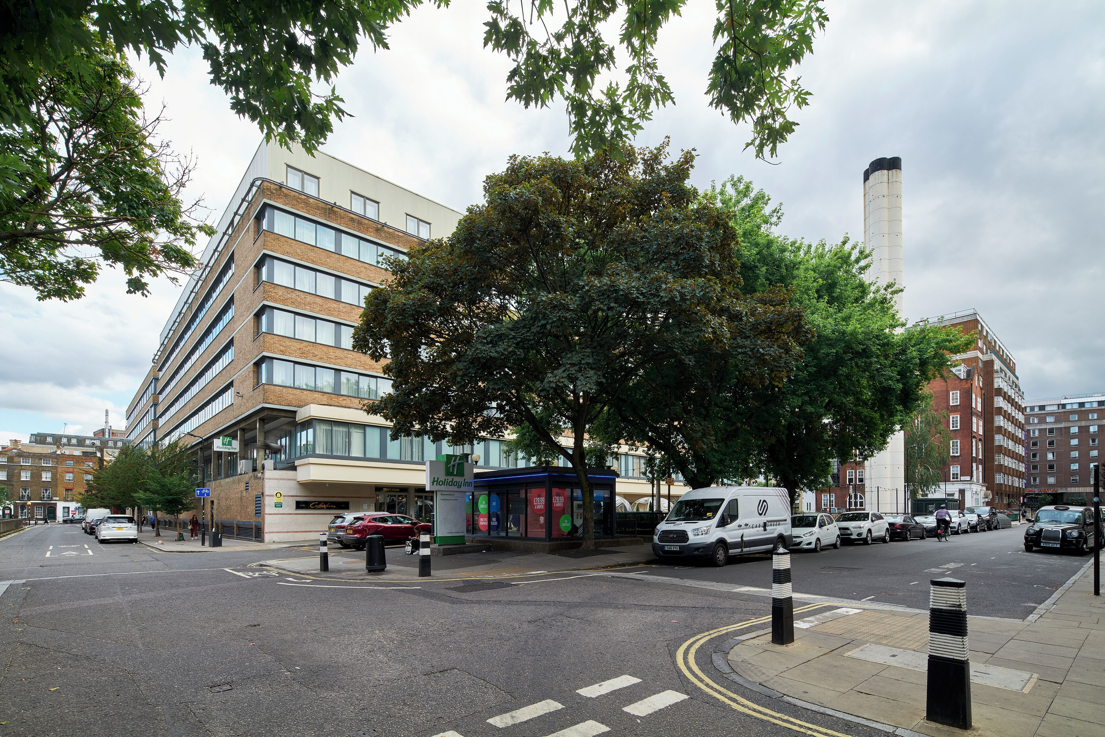A multi-story building with a modern design, a bus stop, and a large tree in front.