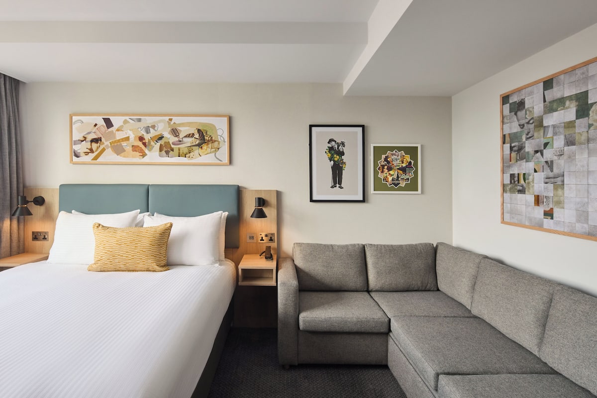 A hotel room with a bed, a grey sofa, and artwork on the walls.