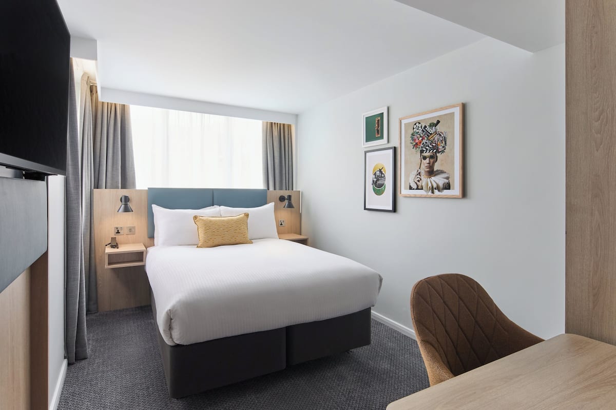 A hotel room with a bed, bedside tables, a TV, a desk, and artwork on the wall.