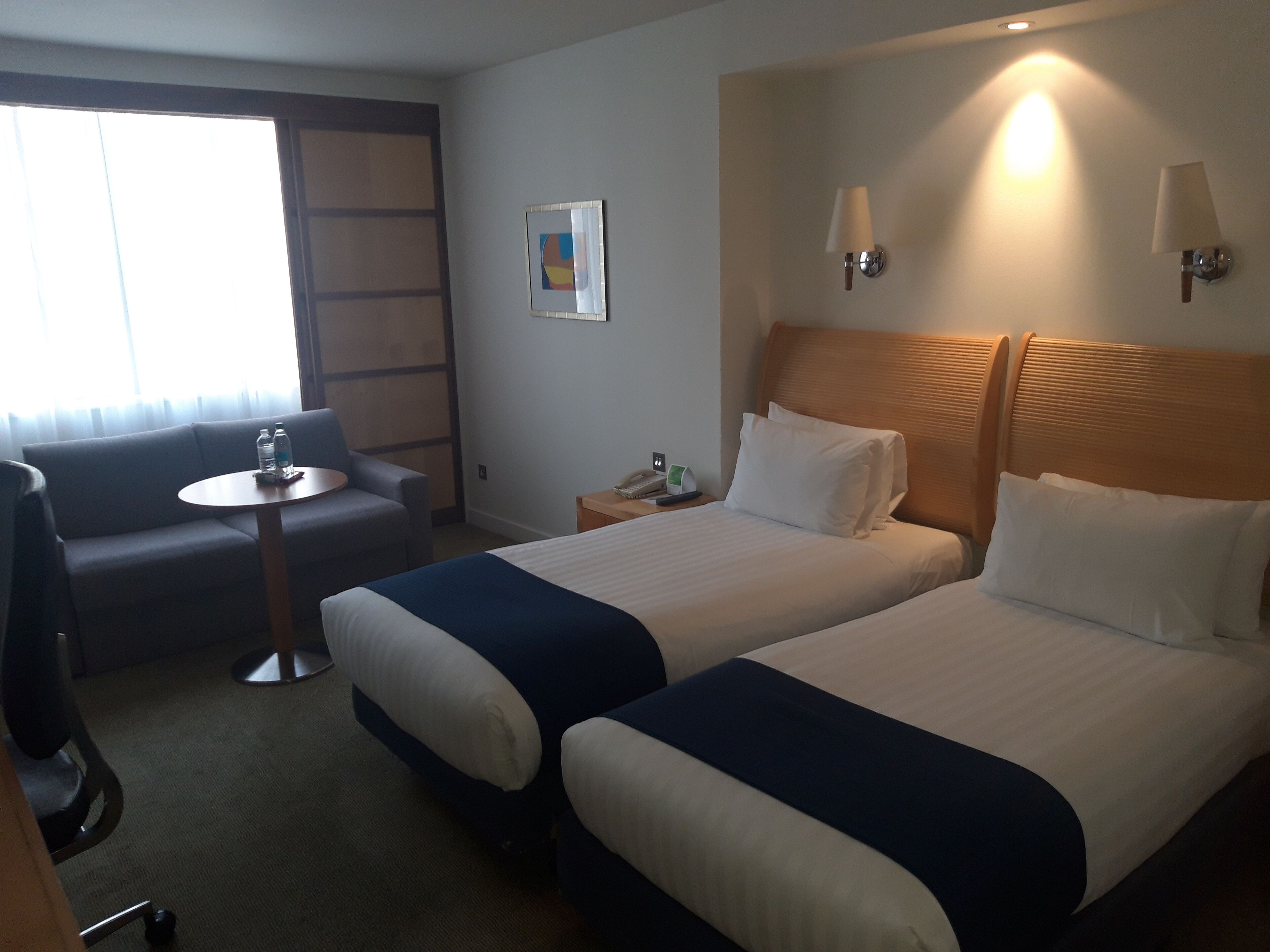 A hotel room with two beds, a sofa, a small table, and a chair. There are wall-mounted lamps and a painting on the wall.
