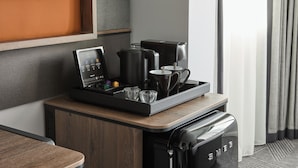 Une chambre d’hôtel moderne avec un coin café sur un comptoir en bois, un téléviseur fixé au mur et une vue sur l’extérieur par une fenêtre avec des rideaux.
