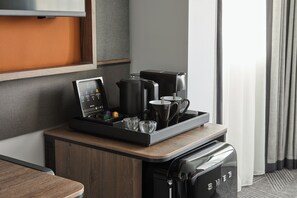 Habitación de hotel moderna con estación de café sobre un mostrador de madera, un televisor montado en la pared y vistas al exterior a través de una ventana con cortinas.