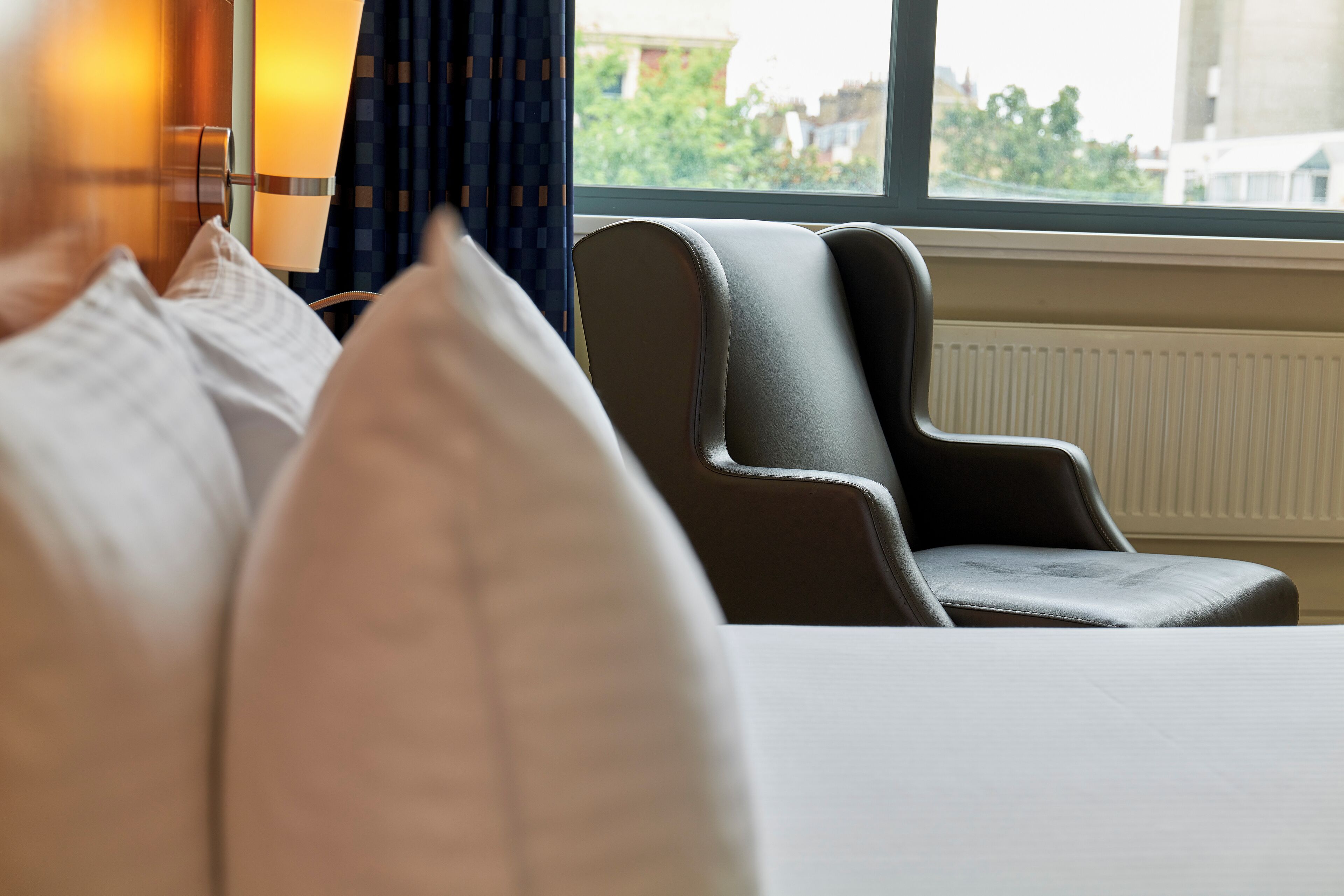 Habitación de hotel con cama, dos sillones, una lámpara y vistas al exterior por una ventana.