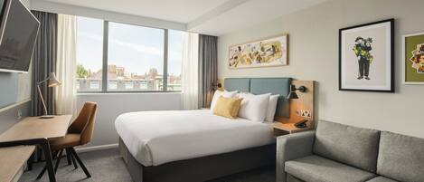 A hotel room with a bed, a desk, a chair, a sofa, and a large window offering a city view.