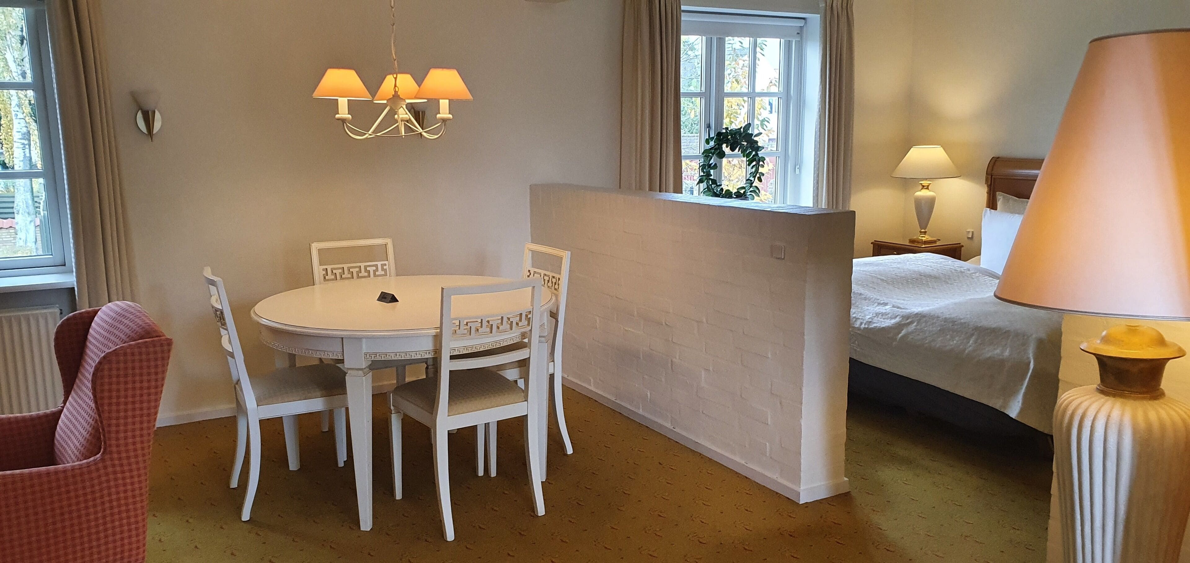 Una habitación de hotel con una mesa de comedor redonda, sillas blancas, una cama con edredón blanco, una mesita de noche con lámpara y una ventana con cortinas.