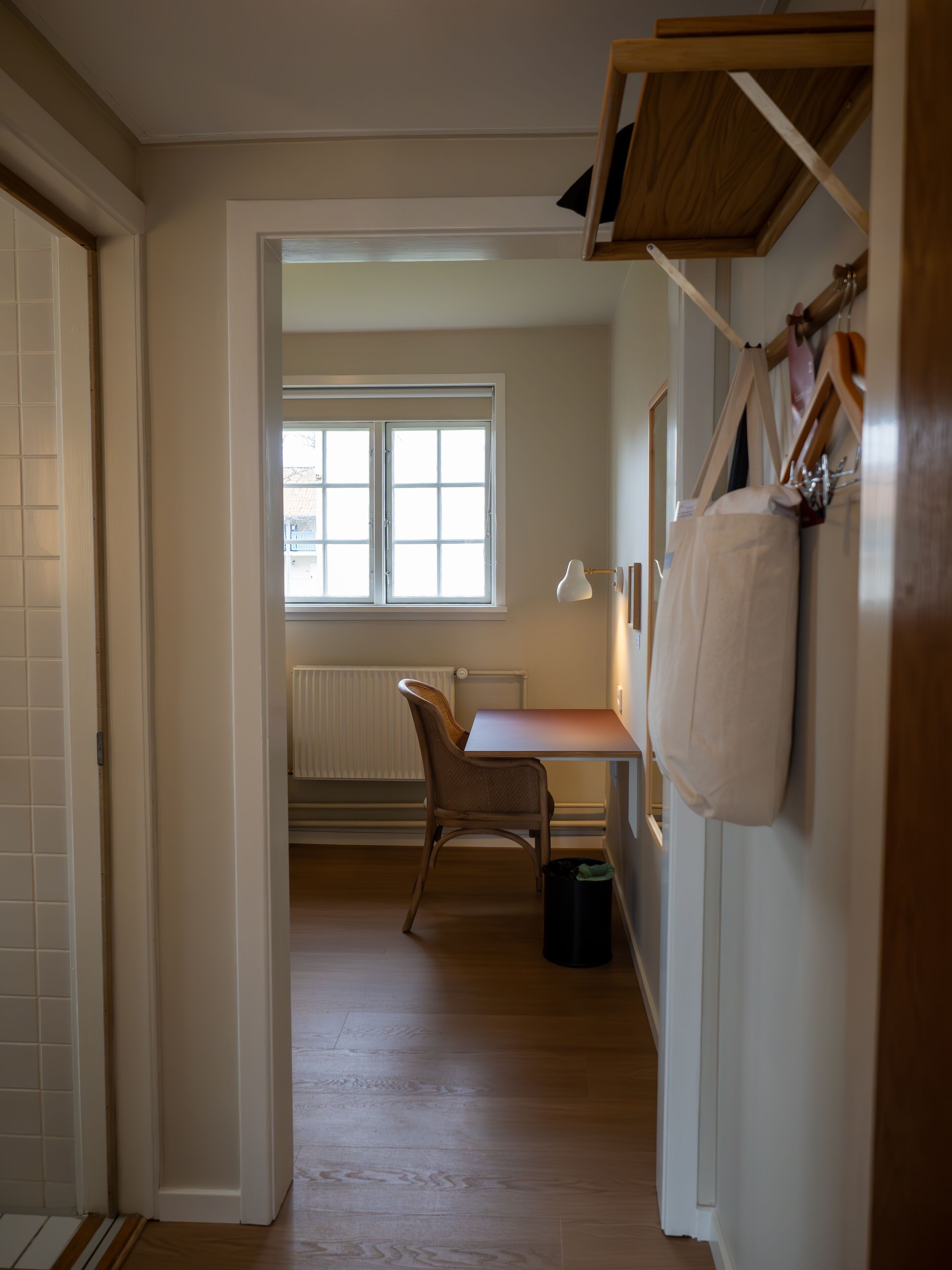 A hallway leading to a room with a wooden floor, a radiator, a window, and a desk with a chair.