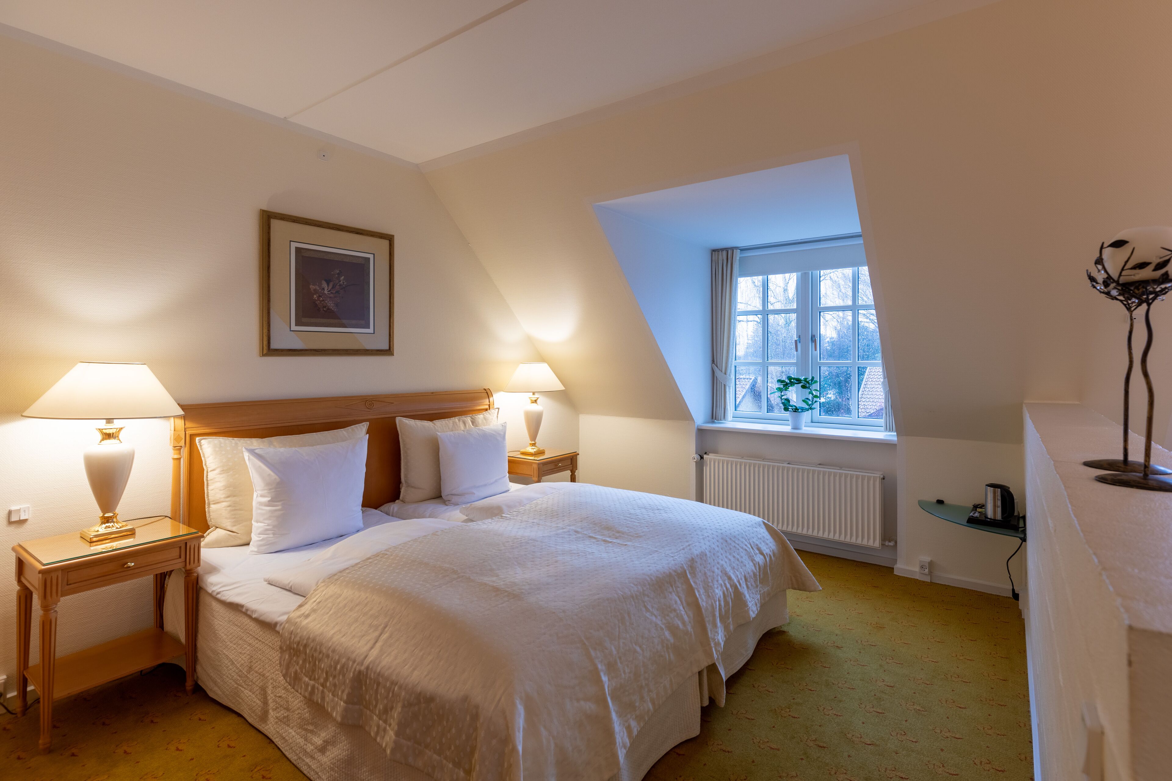A neatly arranged bedroom with a large bed, two bedside tables with lamps, a framed picture on the wall, a window with a view, and a radiator.