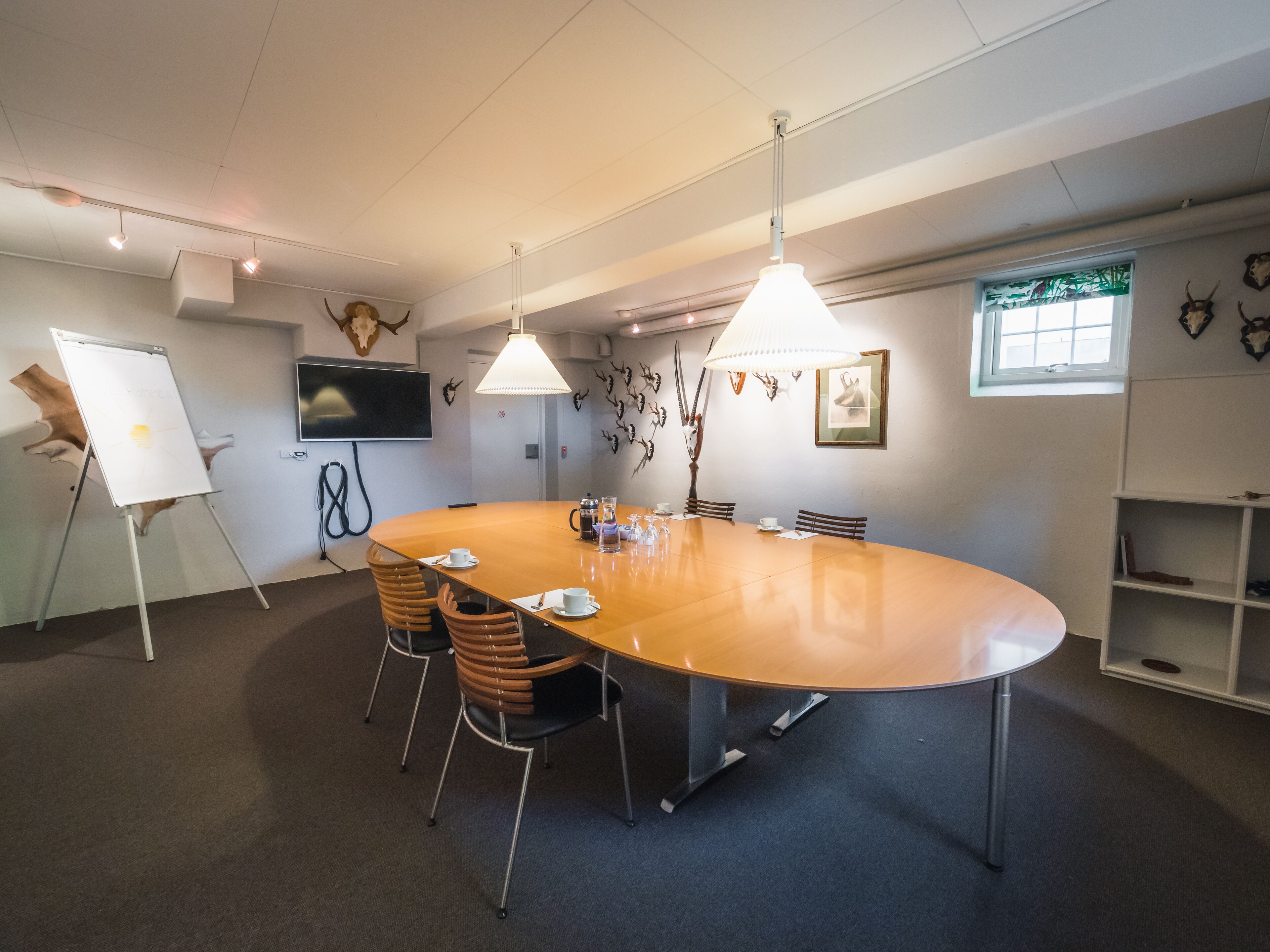 Un salón de conferencias con una gran mesa ovalada, cuatro sillas, una pizarra, un televisor y elementos decorativos en la pared.