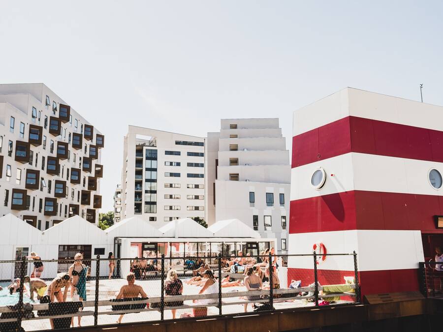 A swimming pool area with people, a large building with a red and white facade, and modern apartment buildings in the background.