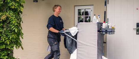 A person pushing a cleaning cart with various cleaning supplies and a white bag.
