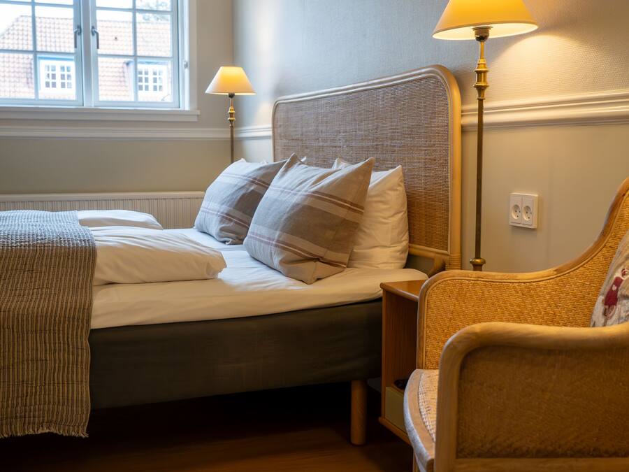 A bedroom with a bed, two bedside lamps, a chair, and a window with a view of buildings.