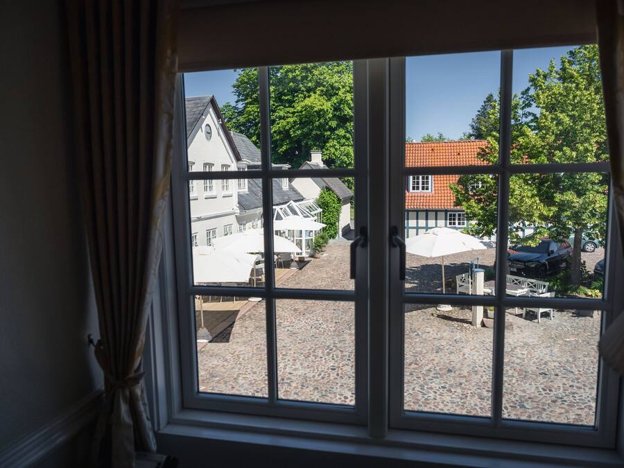 A window with a view of a cobblestone street, buildings, and trees.