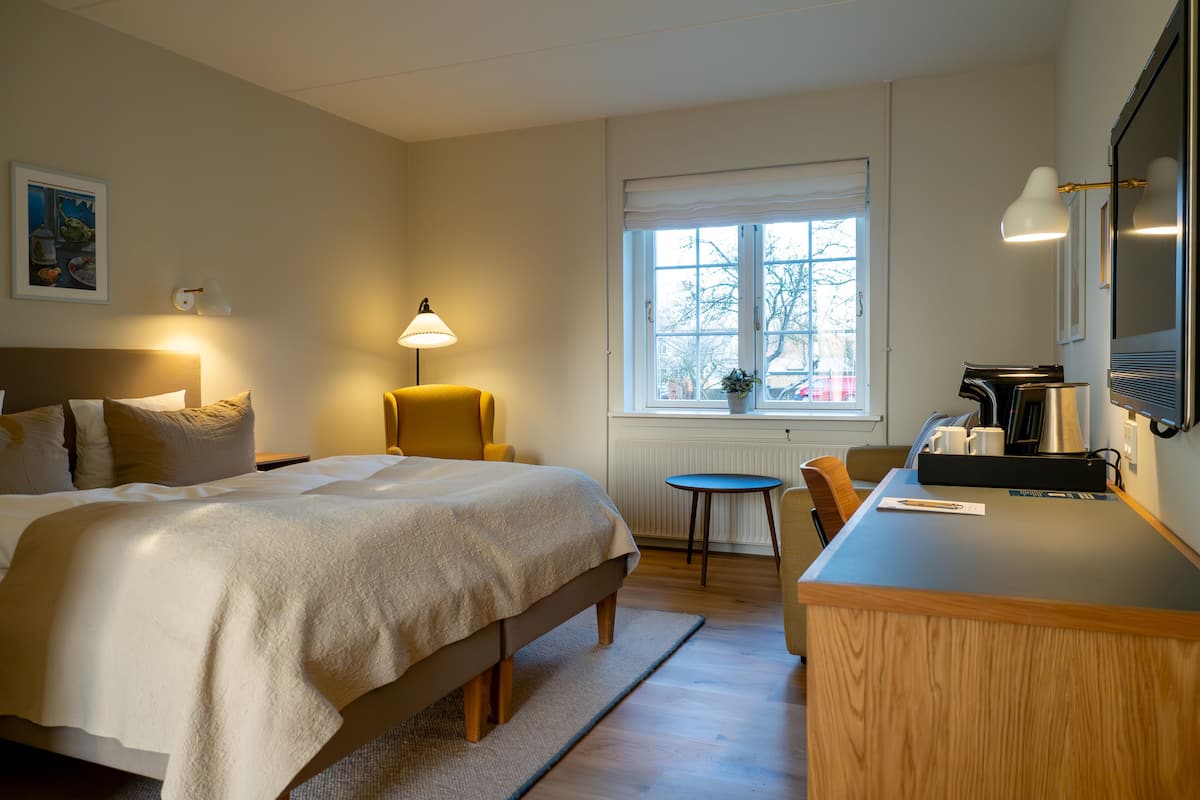 A hotel room with a bed, a desk with a lamp, a chair, and a window with a view of the outside.