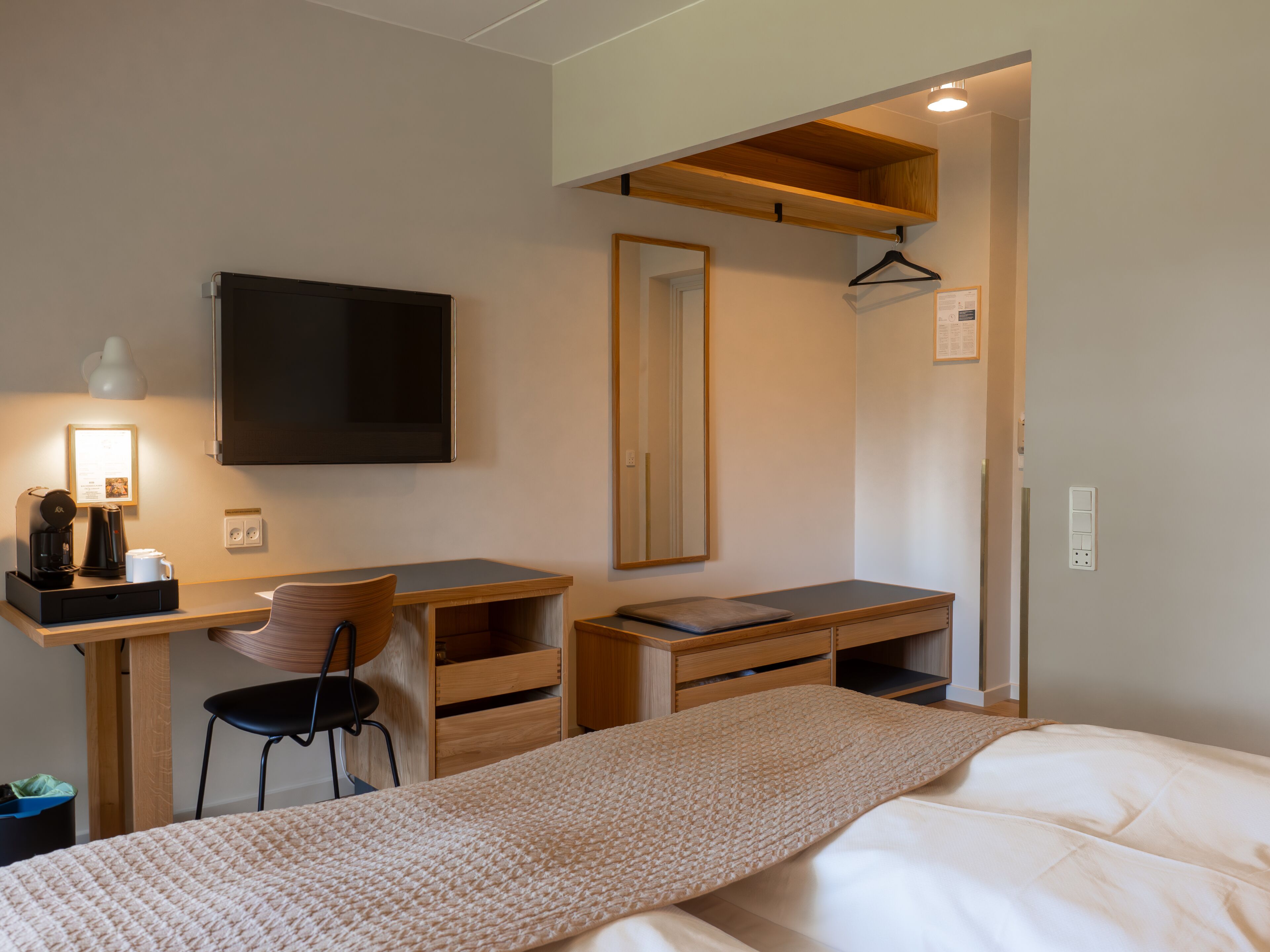 A modern hotel room with a bed, a desk with a chair, a flat-screen TV, a mirror, and a wooden shelf.