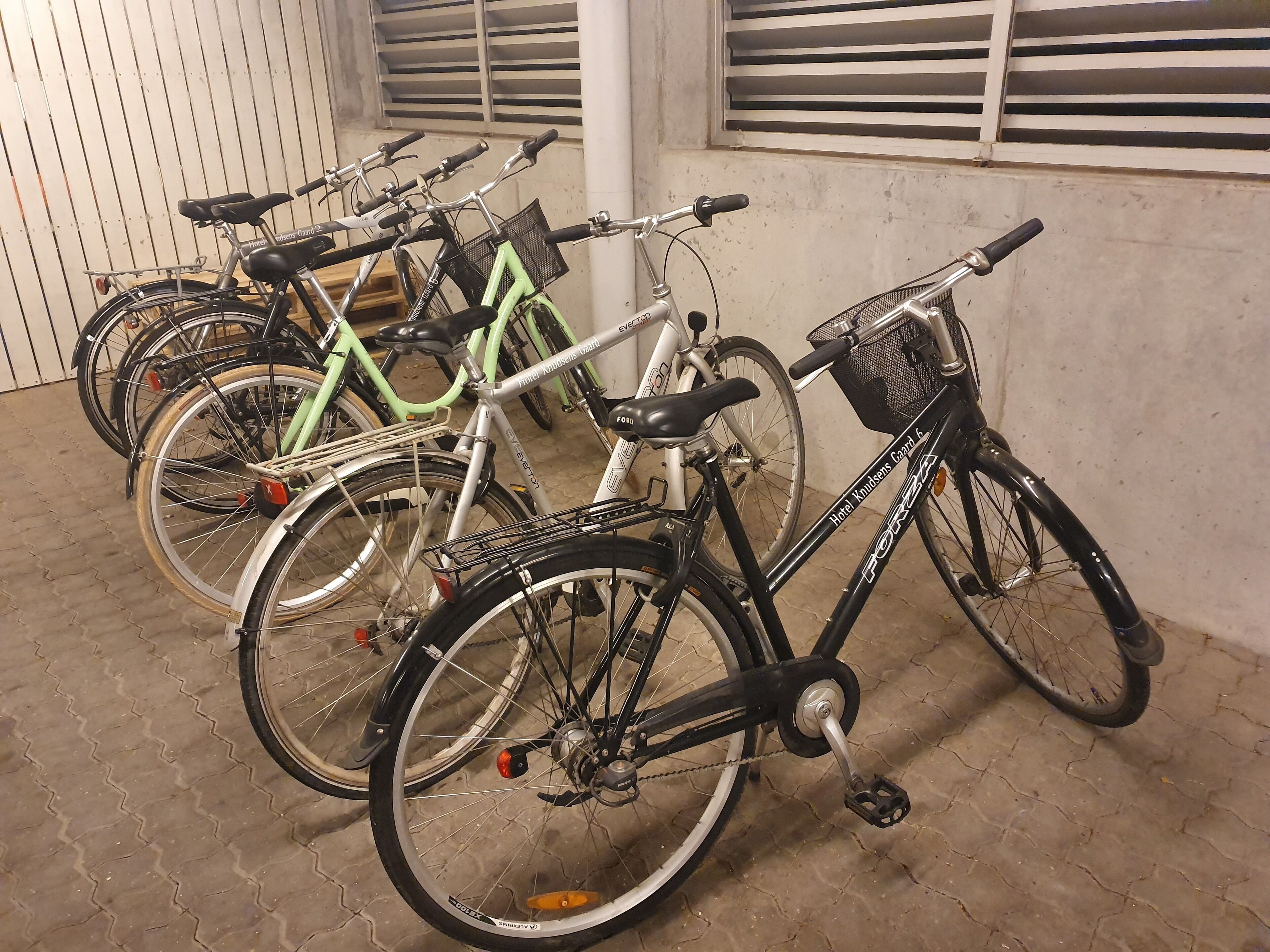 Bicycles parked indoors, with a concrete floor and a window with blinds in the background.