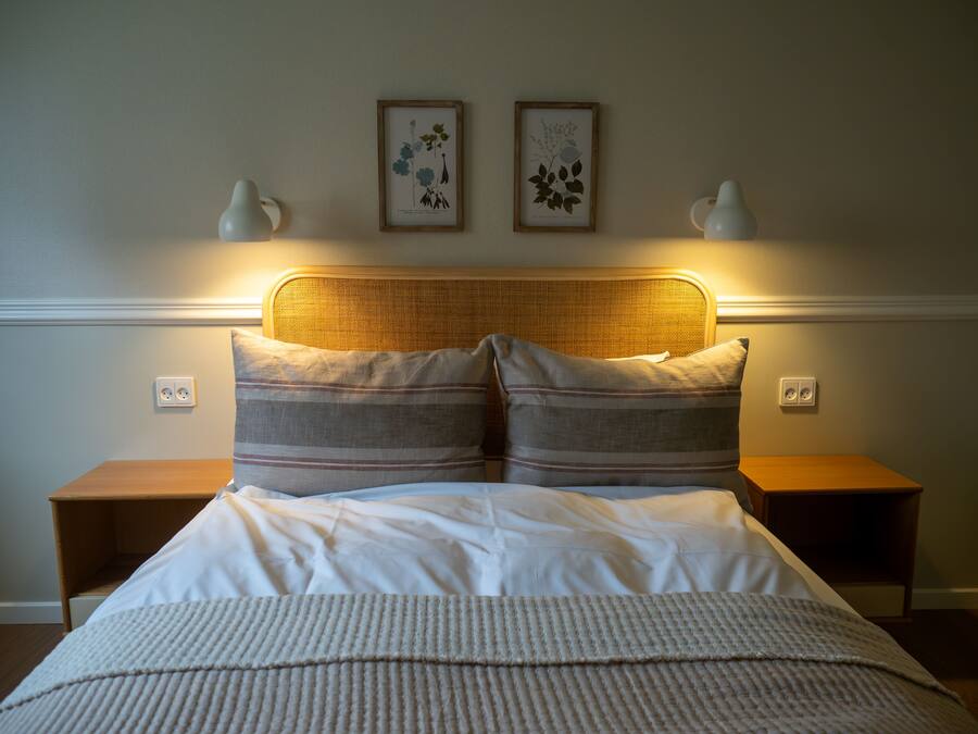 Una cama bien hecha con un almohadón a rayas y una manta con textura, flanqueada por dos mesitas de noche con lámparas, frente a una pared con dos impresiones botánicas enmarcadas.
