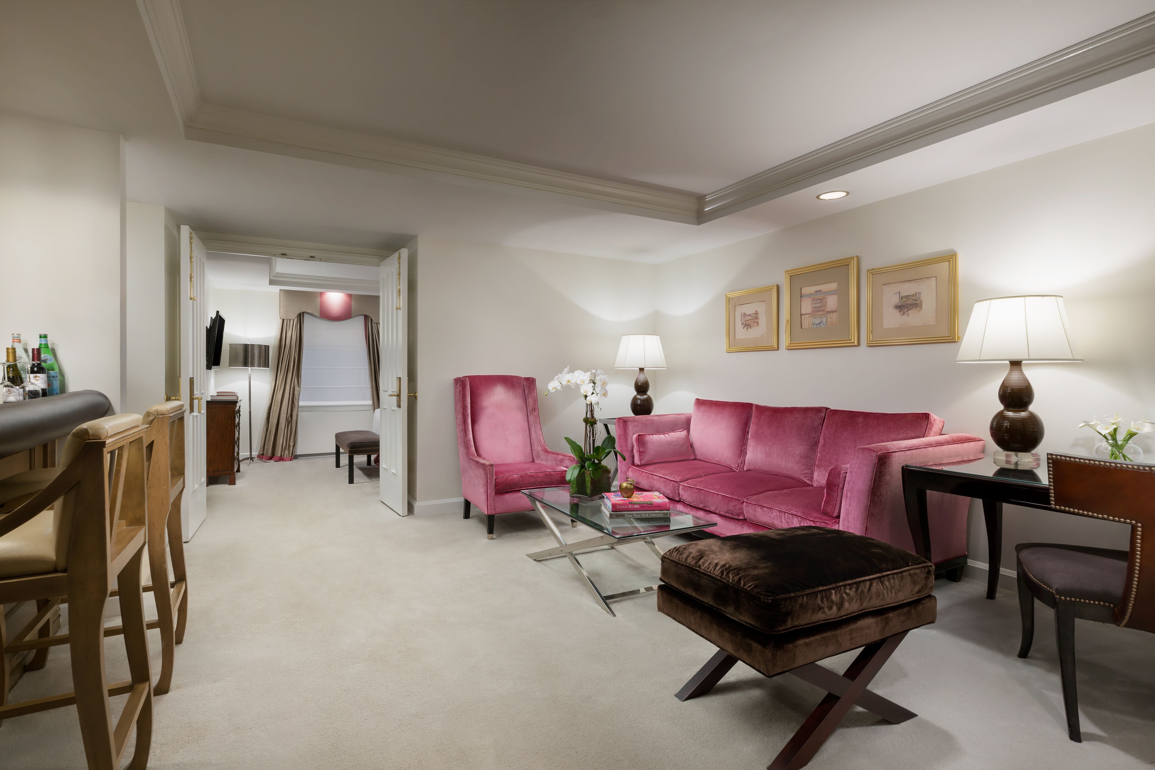 Un salon moderne avec un canapé rose, une table basse en verre et un pouf marron. On y trouve deux fauteuils, une petite table avec une lampe et des tableaux encadrés accrochés au mur.