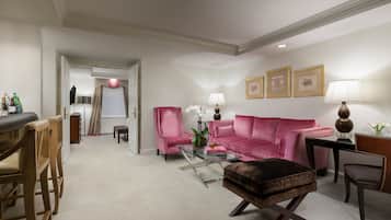 Un salon moderne avec un canapé rose, une table basse en verre et un pouf marron. On y trouve deux fauteuils, une petite table avec une lampe et des tableaux encadrés accrochés au mur.