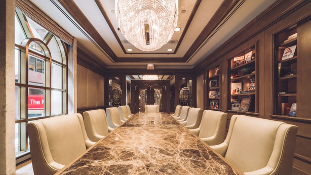 A long, marble-topped conference table with leather chairs in a well-lit room with wooden paneling and a large chandelier.