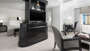 A modern hotel room with a curved black entertainment unit, a bed with white linens, a grey armchair, a small side table with a lamp, and a view of a cityscape through the window.
