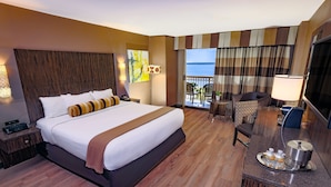 A hotel room with a large bed, a desk with a tray of amenities, a TV, and a balcony with a view of the sea.