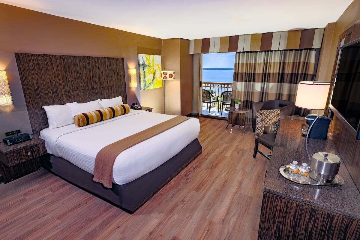 A hotel room with a large bed, a desk with a tray of amenities, a TV, and a balcony with a view of the sea.