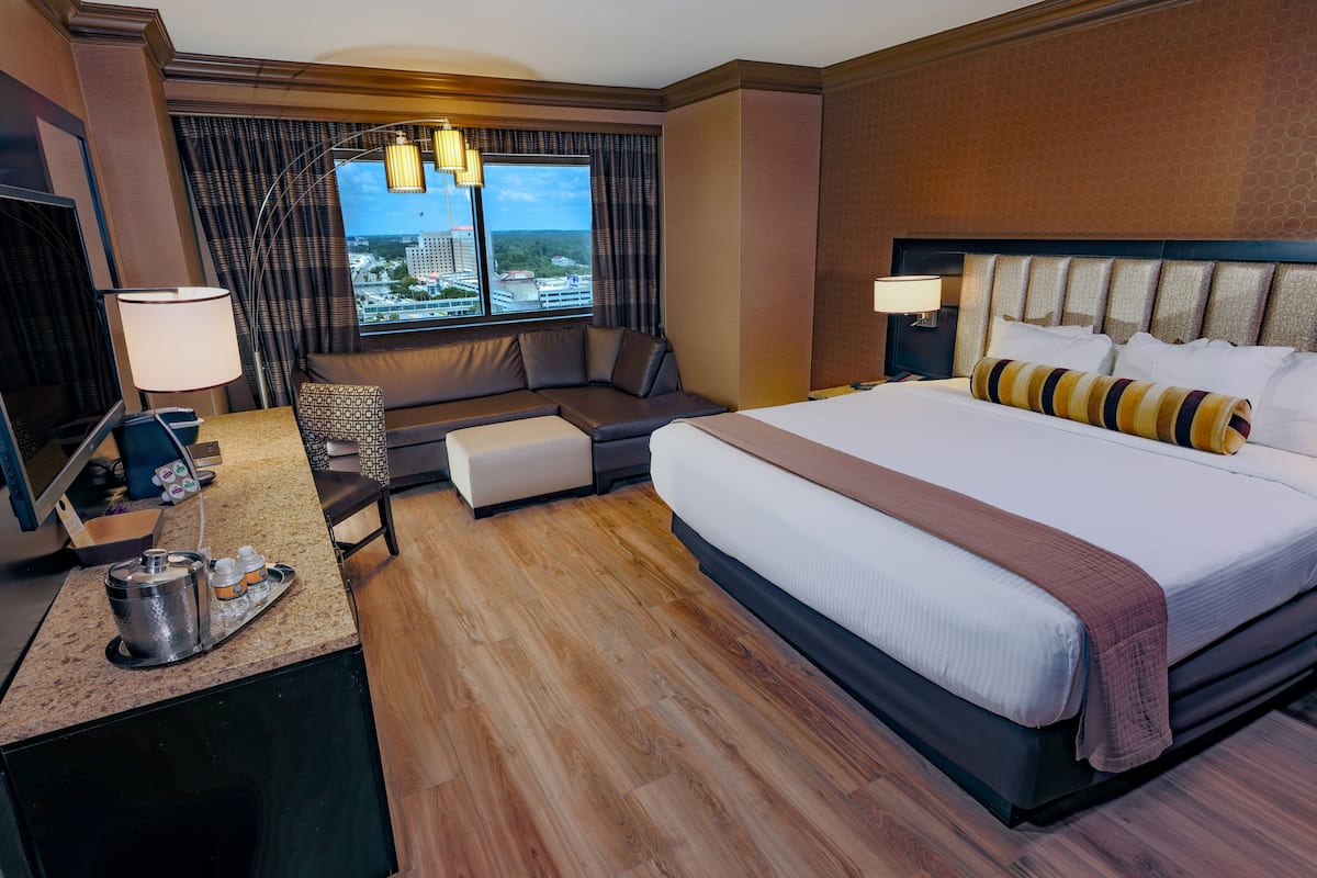 A hotel room with a large bed, a desk with a lamp, a seating area with a view of the city, and a minibar.