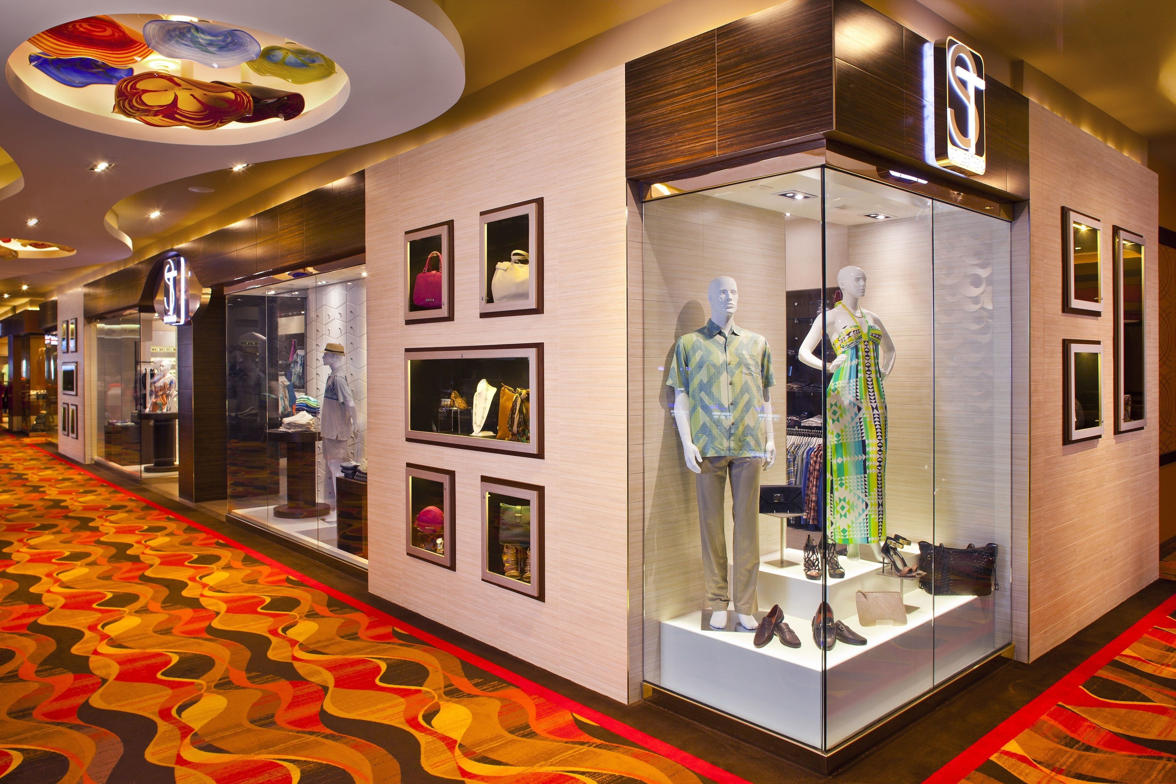 A corridor with a patterned carpet, glass storefronts displaying clothing and accessories, and a ceiling with circular light fixtures.