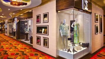 A corridor with a patterned carpet, glass storefronts displaying clothing and accessories, and a ceiling with circular light fixtures.