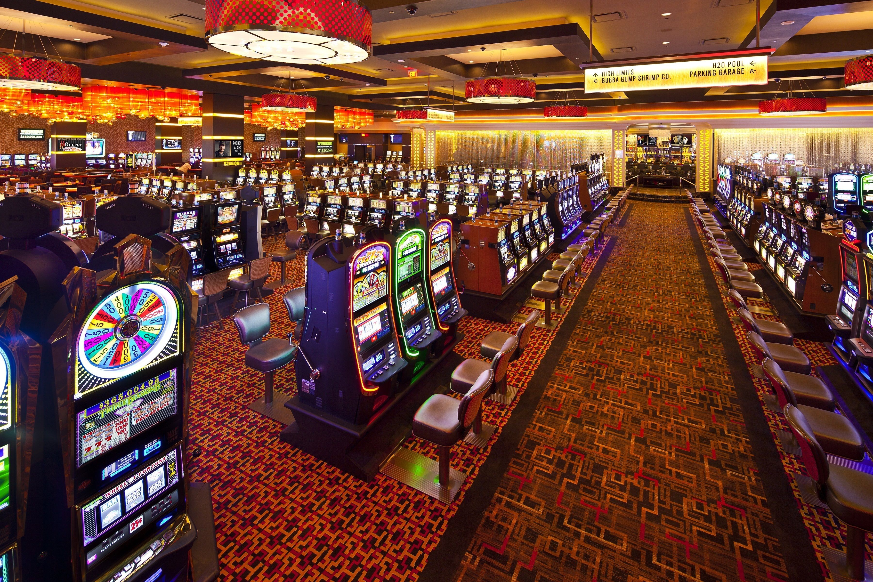 A casino floor with multiple slot machines, a carpeted floor, and a bar area in the background.