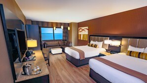 A hotel room with two beds, a desk with a TV, a seating area, and a view of the sea.