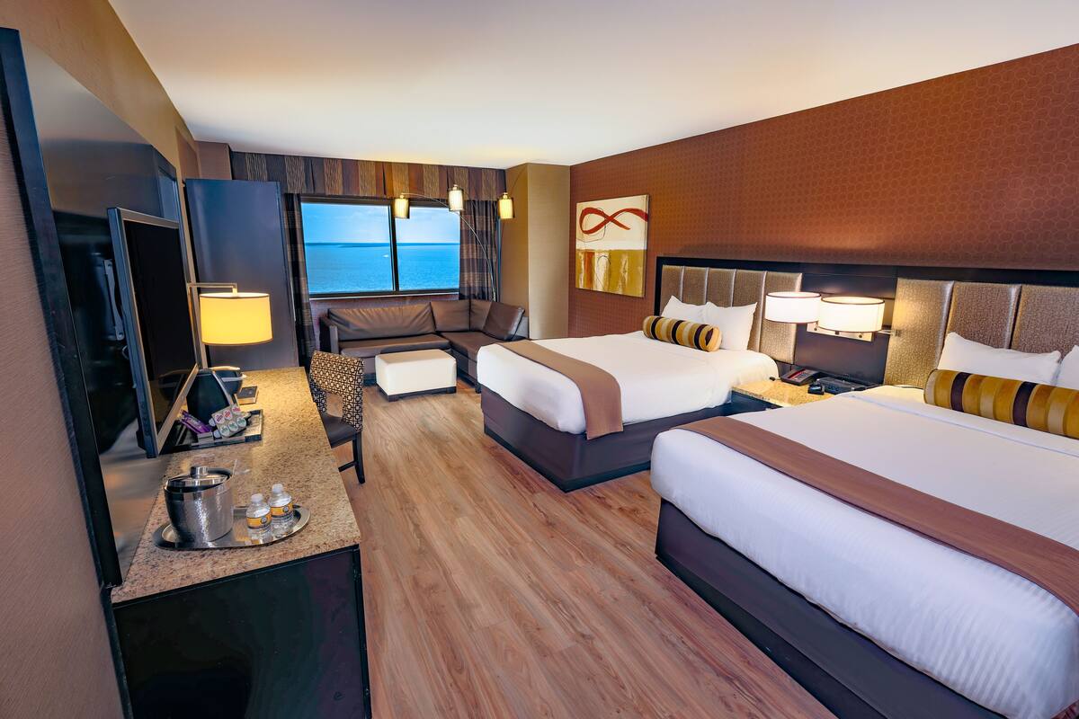 A hotel room with two beds, a desk with a TV, a seating area, and a view of the sea.