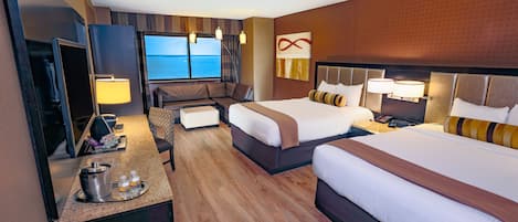 A hotel room with two beds, a desk with a TV, a seating area, and a view of the sea.