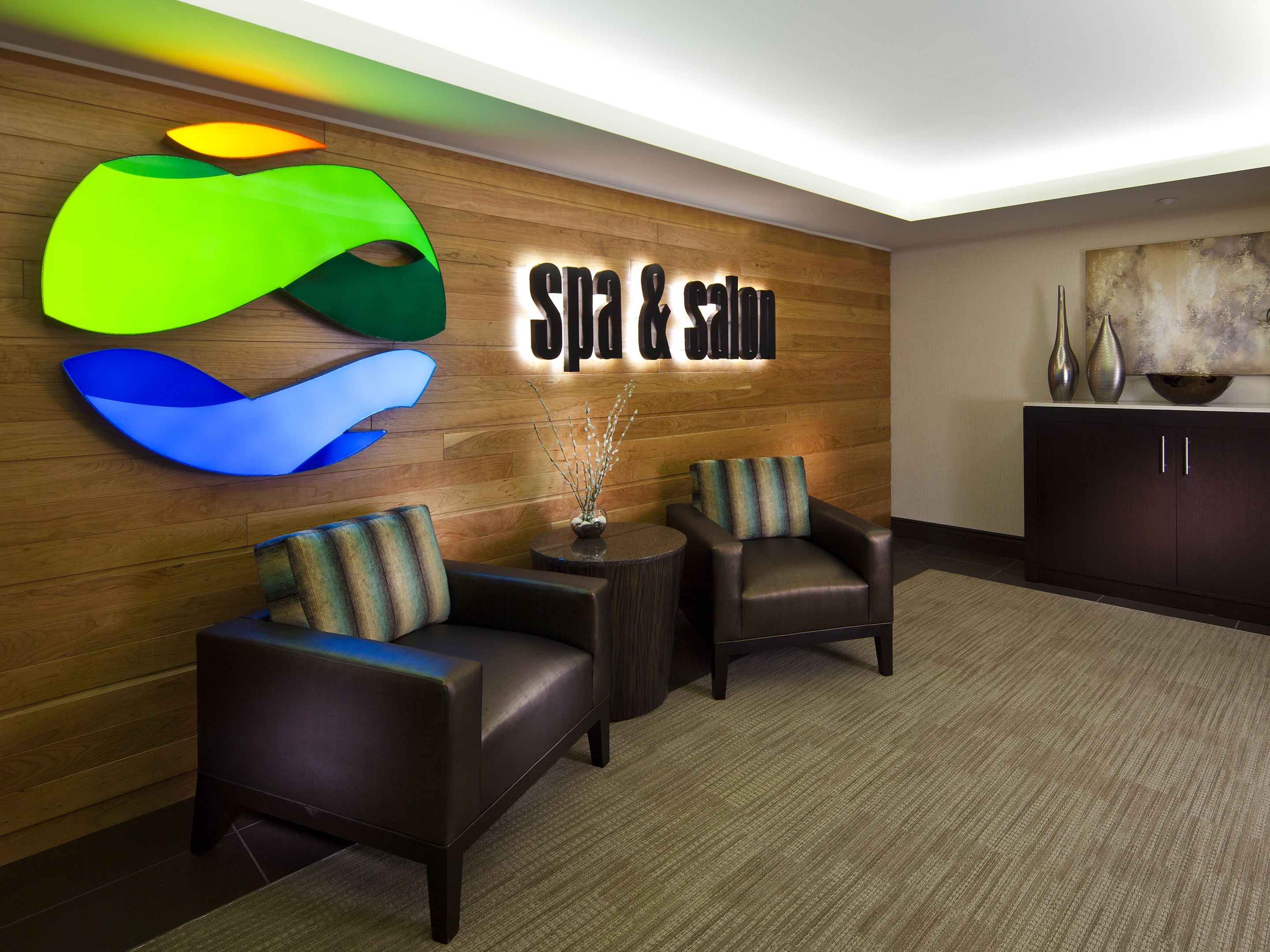 A modern lobby with a 'Spa & Gym' sign, leather armchairs, a small table with a vase, and a cabinet with decorative vases.