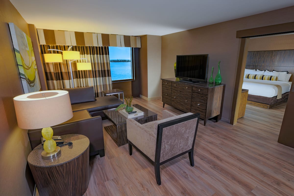 A hotel room with a sofa, a coffee table, a TV, a bed, and a view of the water.