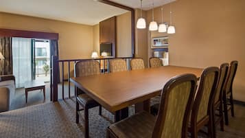 A hotel dining area with a large wooden table, six upholstered chairs, a television, and a balcony with a view.