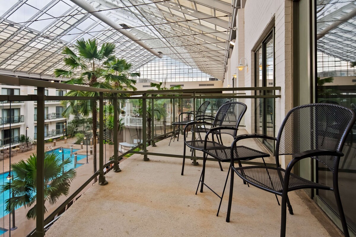 A balcony with a glass ceiling overlooking a pool and a building with multiple floors.