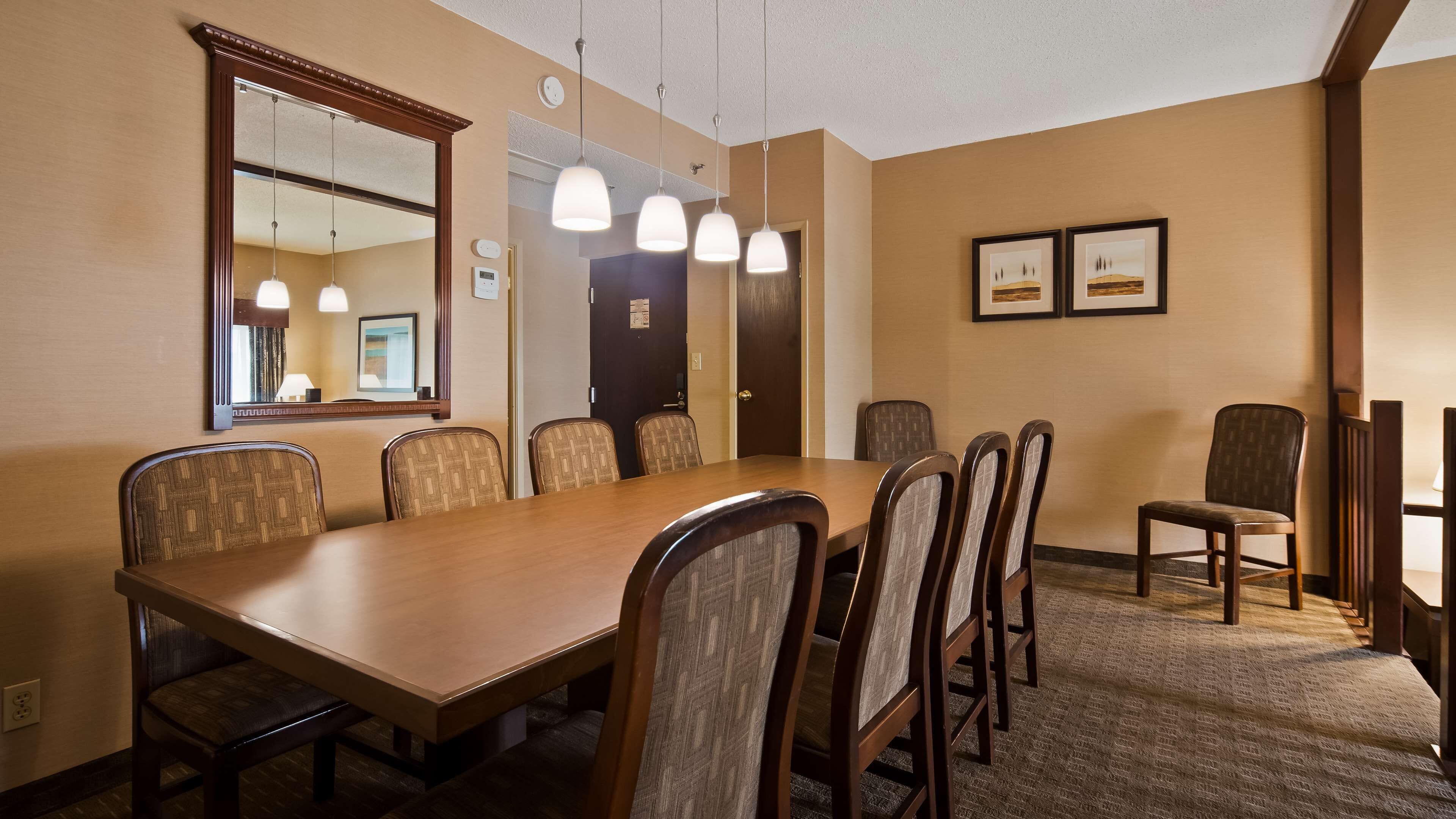 A conference room with a long table, chairs, pendant lights, a large mirror, framed artwork, and a door leading to another room.
