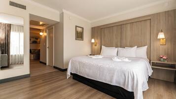 Une chambre d’hôtel avec un grand lit, une tête de lit en bois, deux lampes de chevet, une petite table avec un vase et une vue sur la chambre adjacente.