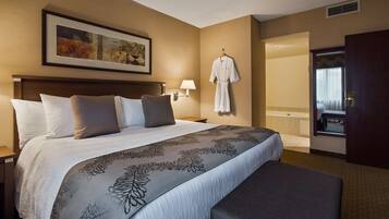 Quarto de hotel com uma cama grande, uma obra de arte emoldurada acima da cama, um roupão pendurado na parede e vista para o banheiro por uma porta aberta.