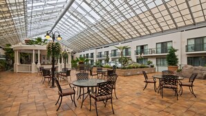 A spacious indoor patio with multiple tables and chairs, a glass ceiling, and various plants and decorative elements.