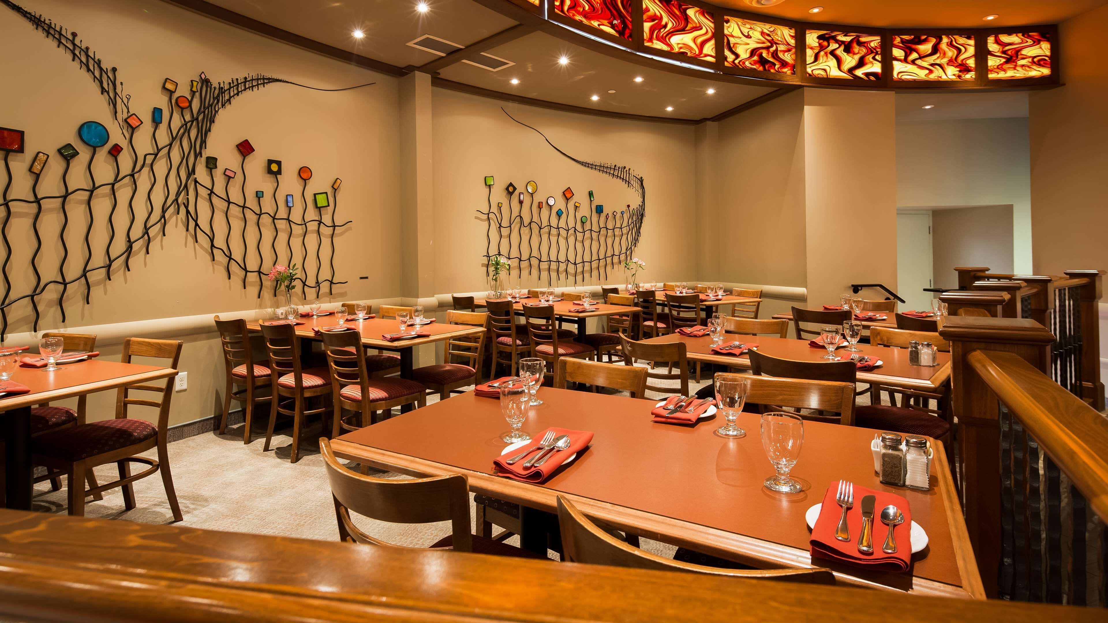 A restaurant with multiple tables and chairs, a decorative wall with abstract art, and a staircase leading to another level.