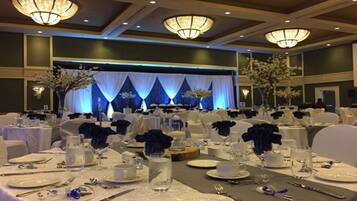 Un salón de banquetes con mesas redondas dispuestas para un evento formal, con manteles blancos, centros de mesa florales azules e iluminación elegante.