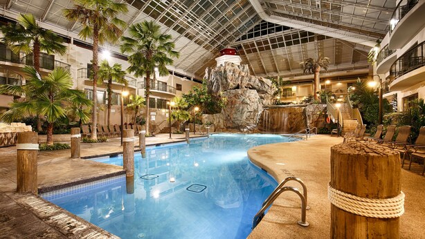 An indoor swimming pool with a waterfall feature, surrounded by lounge chairs and palm trees.