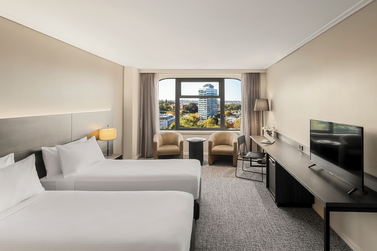A hotel room with two beds, a desk, a TV, and a view of a cityscape.