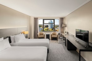 A hotel room with two beds, a desk, a TV, and a view of a cityscape. - The Sydney Boulevard Hotel by Accor (Woolloomooloo)