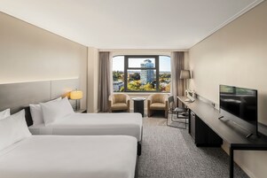 Quarto de hotel com duas camas, uma escrivaninha, uma televisão e vista para a cidade.