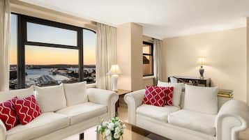 A hotel room with two white sofas, a coffee table, a desk with a lamp, and a view of a cityscape through large windows.